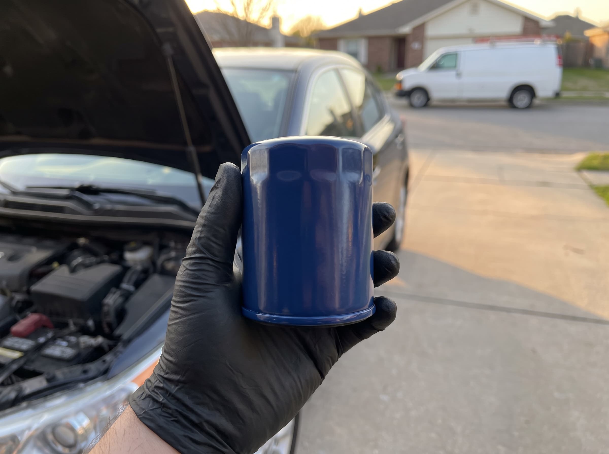 A gloved hand holding a new oil filter beside an open hood in a driveway
